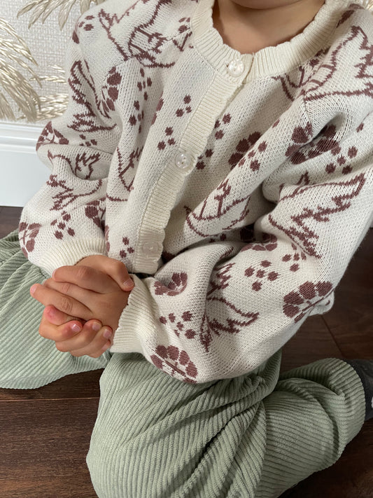 Toddler sits kneeling on the floor with clasped hands, wearing a cream knitted cardigan with a jacquard knit pattern of brown crane birds flying among floral motifs. The cardigan is paired with sage green corduroy trousers.