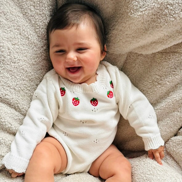 Baby girl wearing a white knitted onesie romper with red strawberry patterns on a soft surface