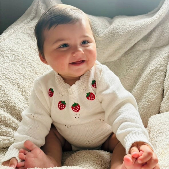 9 month old baby girl wearing white knitted cotton baby romper with strawberry embroidery detail around the neckline. Baby girl looking beyond the camera smiling, holding onto her feet with her hands with legs extended.