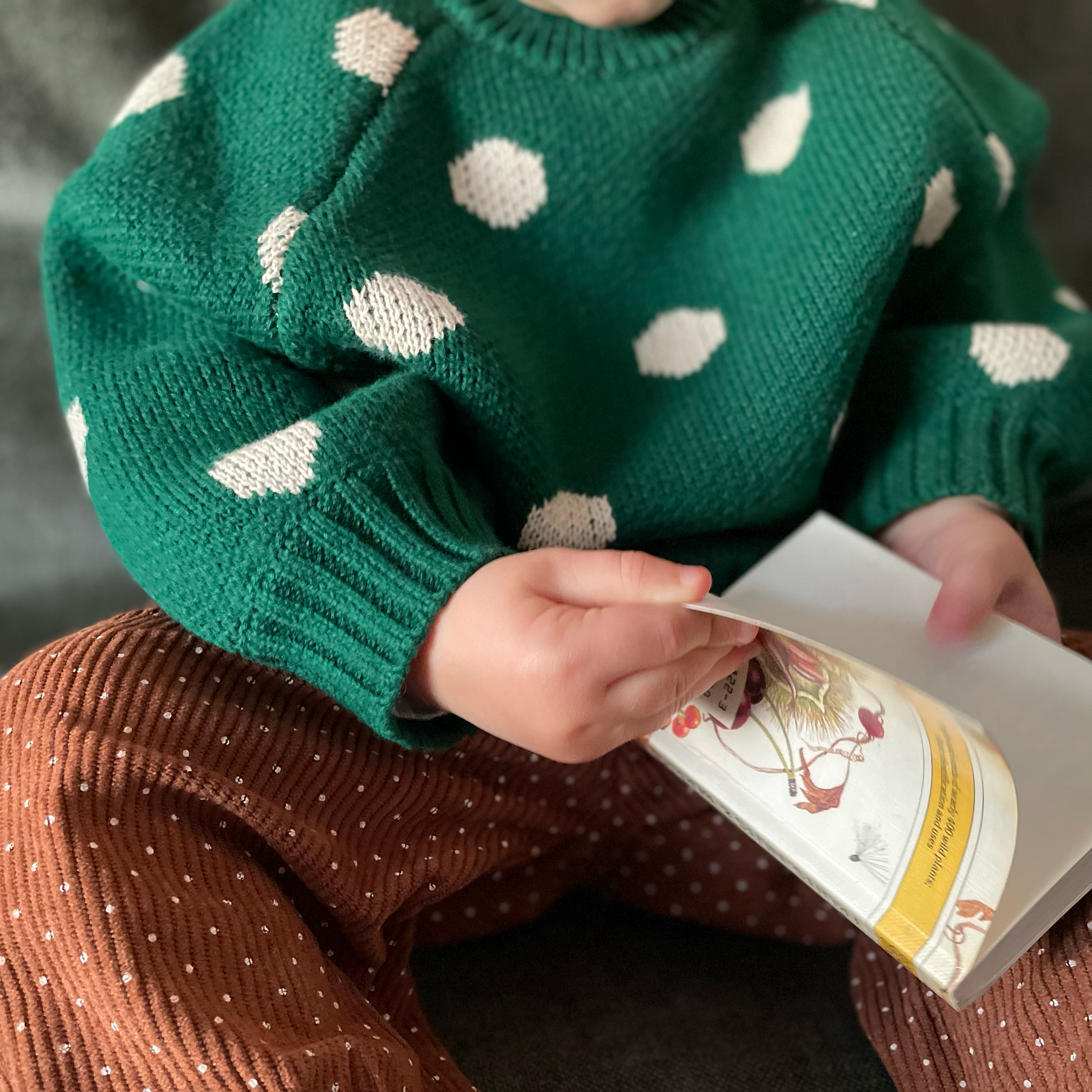 Green knitted jumper with beige jumbo spot pattern worn by toddler reading a book