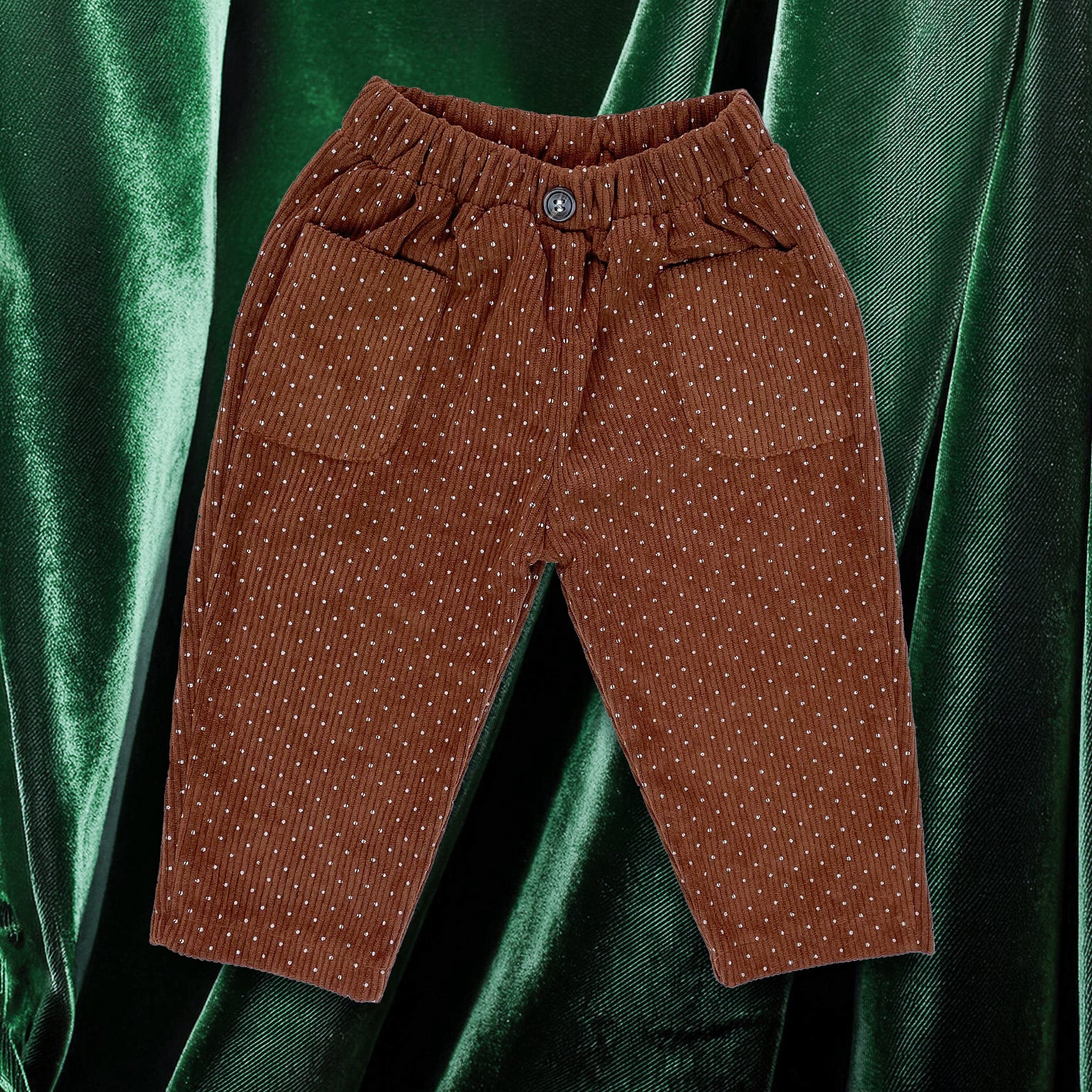 Brown corduroy baby toddler trousers with all over white polka dot pattern. Garment is laid flat on top of a green velvet background
