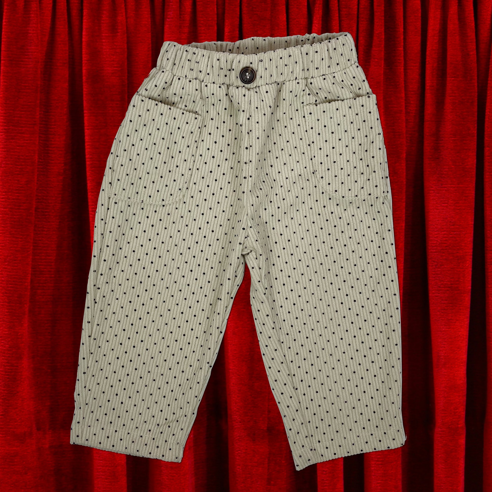 Cream corduroy baby toddler trousers with all over black polka dot print. Trousers are laid flat on a red velvet background.