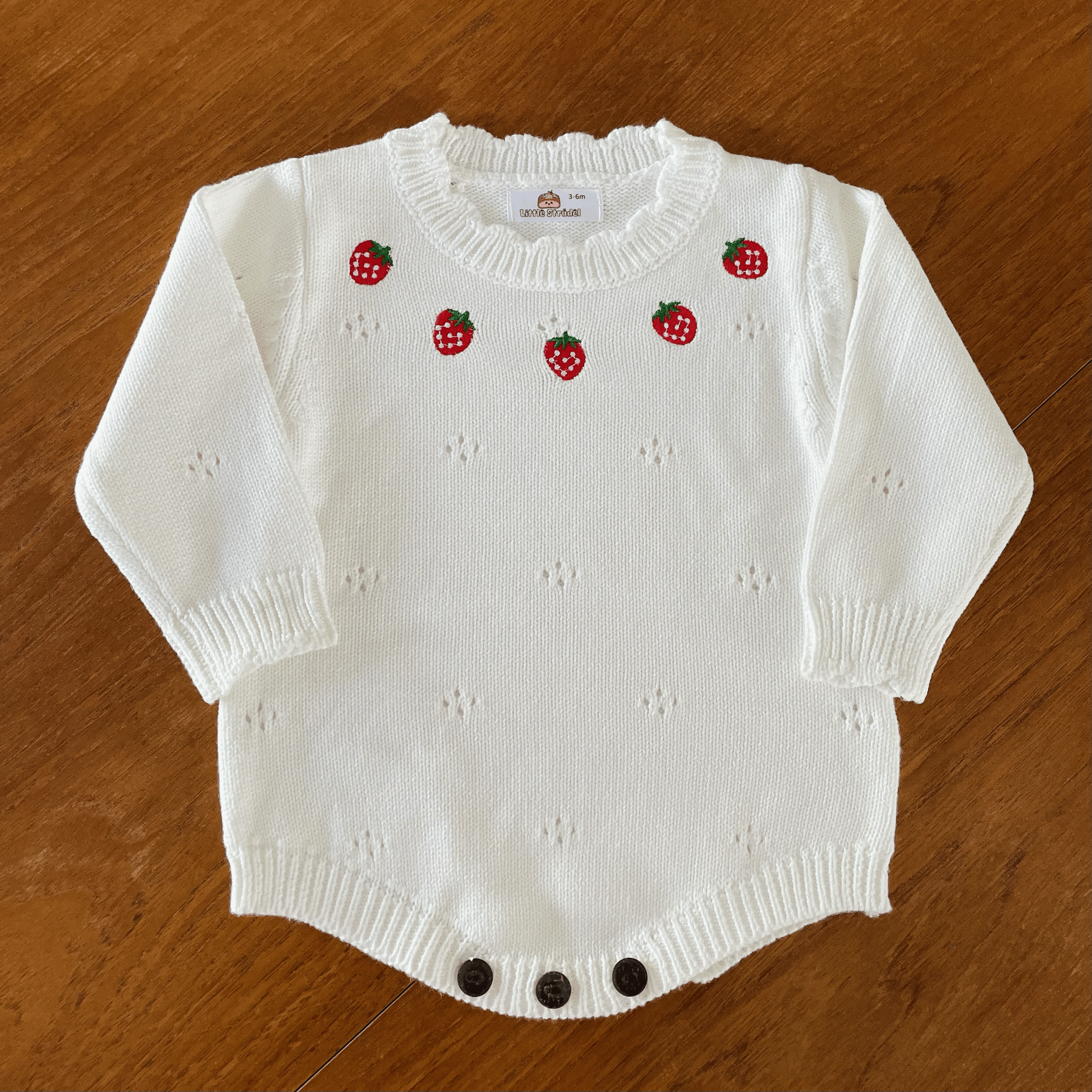 Video of a white knitted baby romper. The camera zooms in to show details of the soft cotton fabric, strawberry embroidery and scallop knit trims. Wooden button fastenings and soft stretchy rib at the leg openings for comfort. 