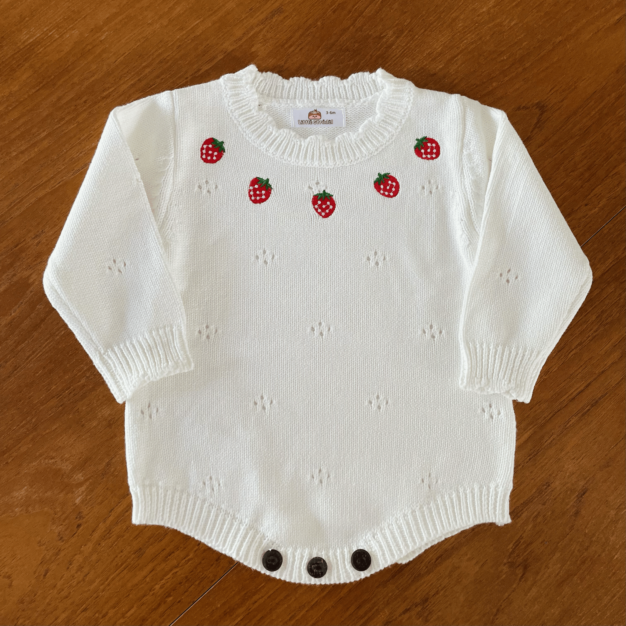 Video of a white knitted baby romper. The camera zooms in to show details of the soft cotton fabric, strawberry embroidery and scallop knit trims. Wooden button fastenings and soft stretchy rib at the leg openings for comfort. 